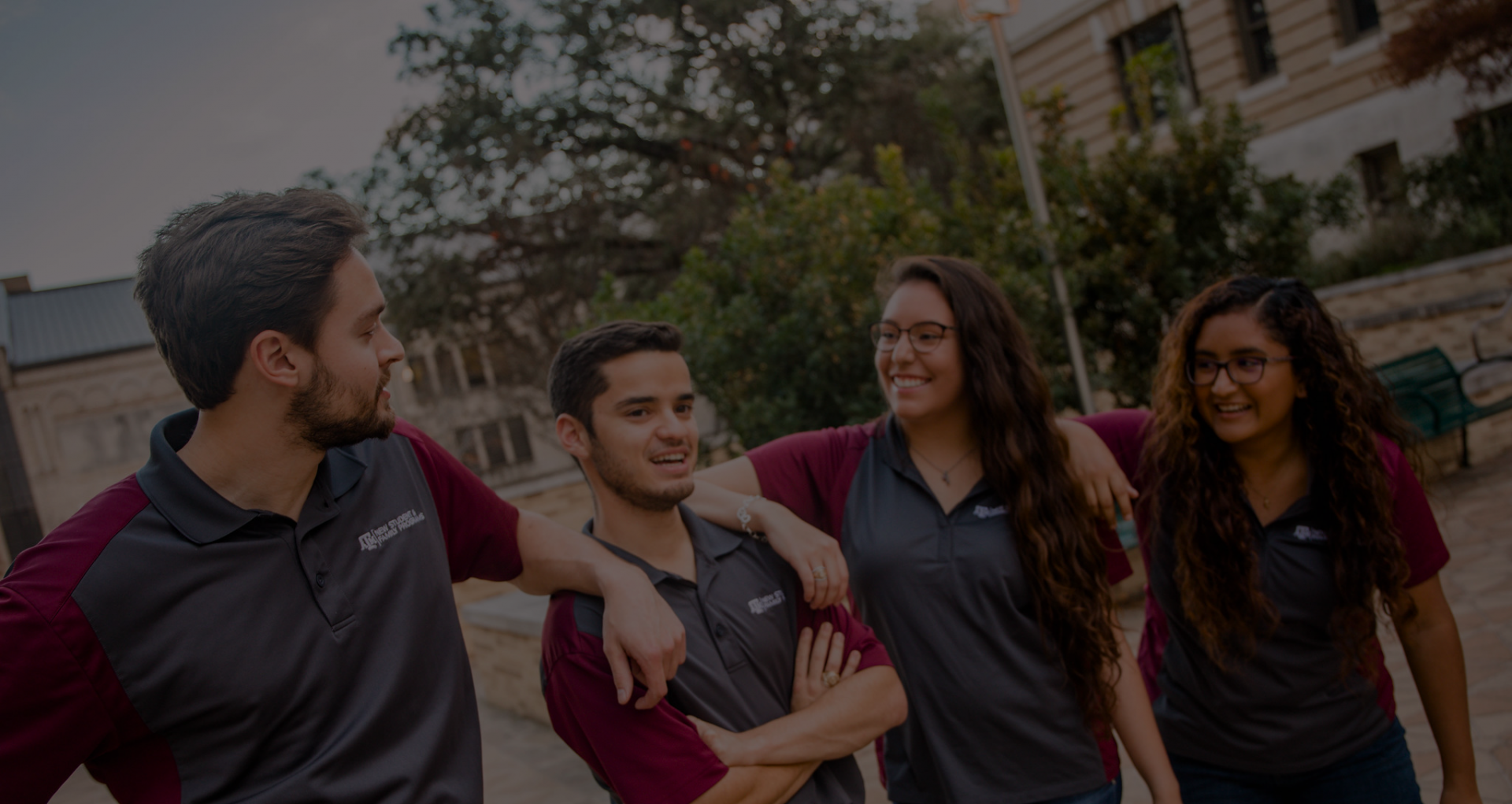 Four student leaders wearing gray and maroon polos stand outside on campus. The photo has a dark gray color overlay.